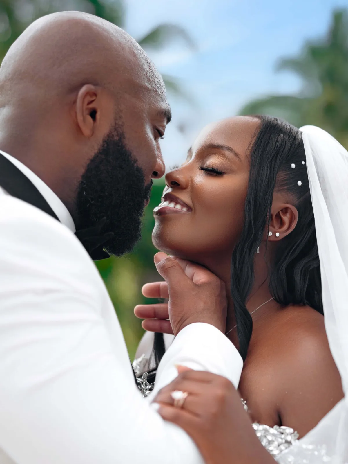 Bald bearded groom in white tuxedo tenderly holding smiling bride in veil outdoors against greenery