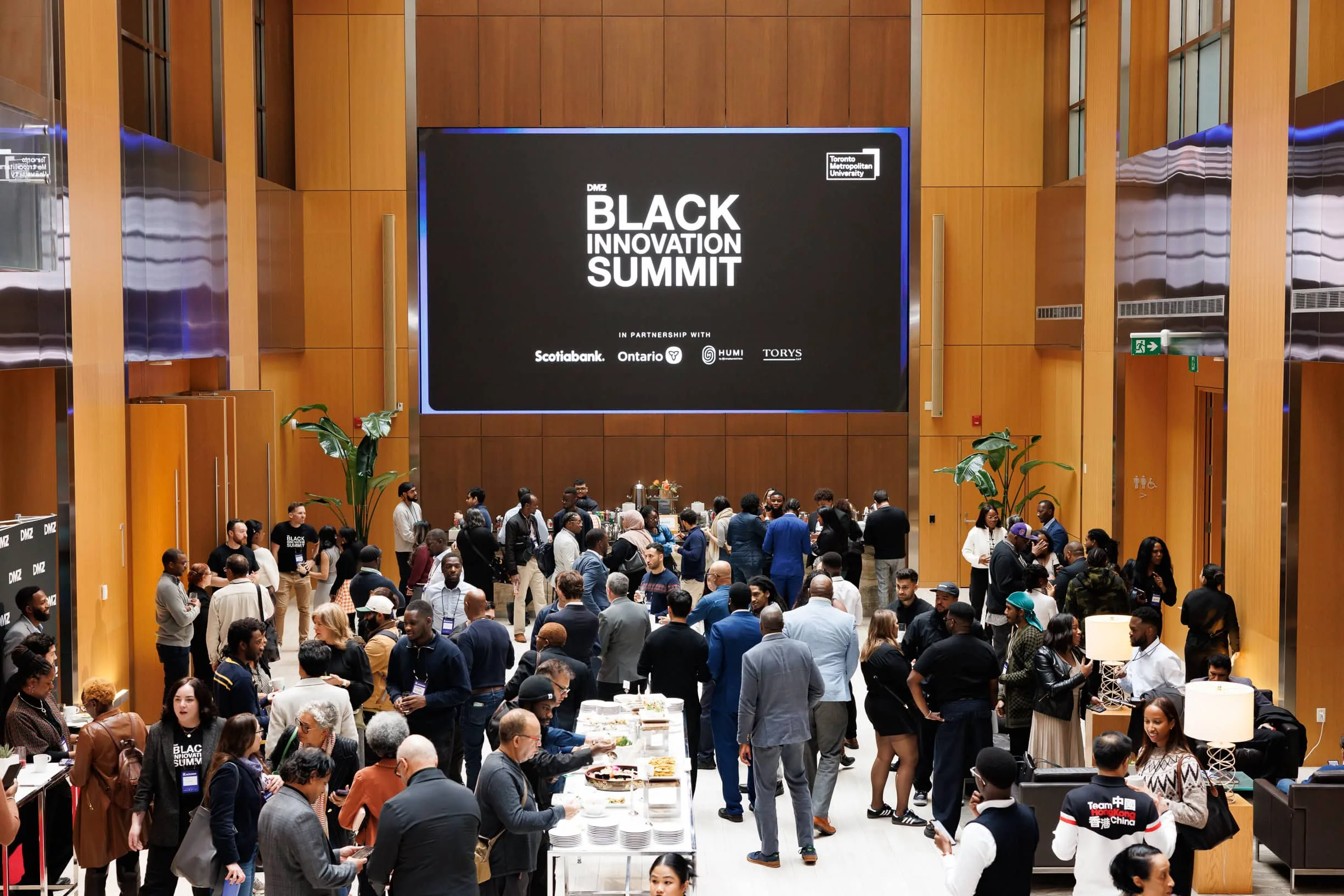 Crowd of professionals mingling at catering tables in wood-paneled conference atrium with large screen