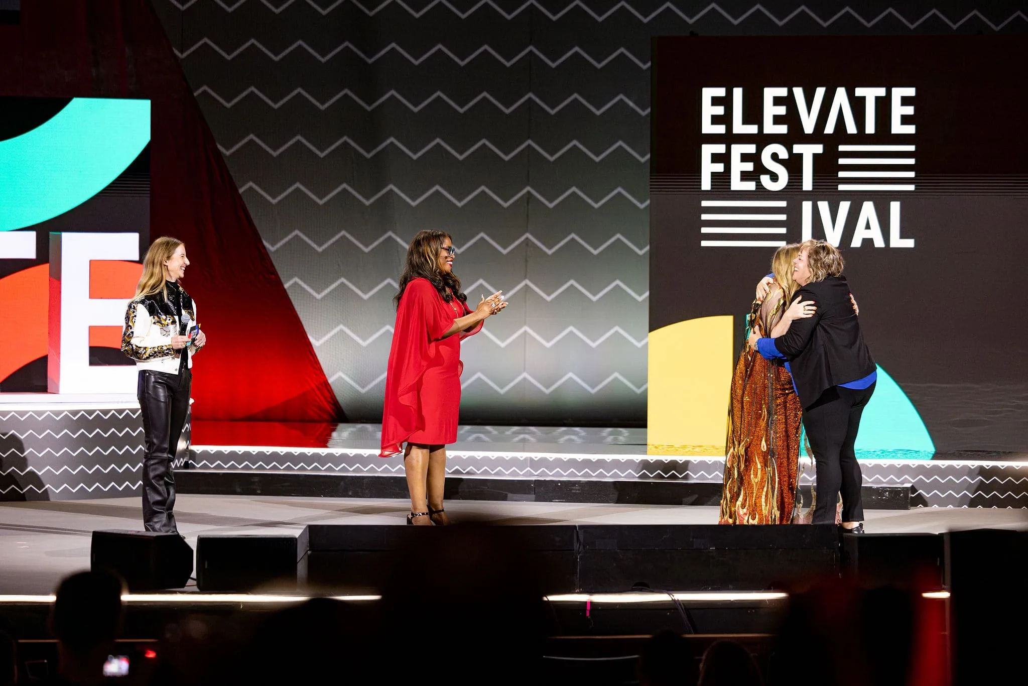 Four women on a colorful event stage; two embrace warmly while another in red claps