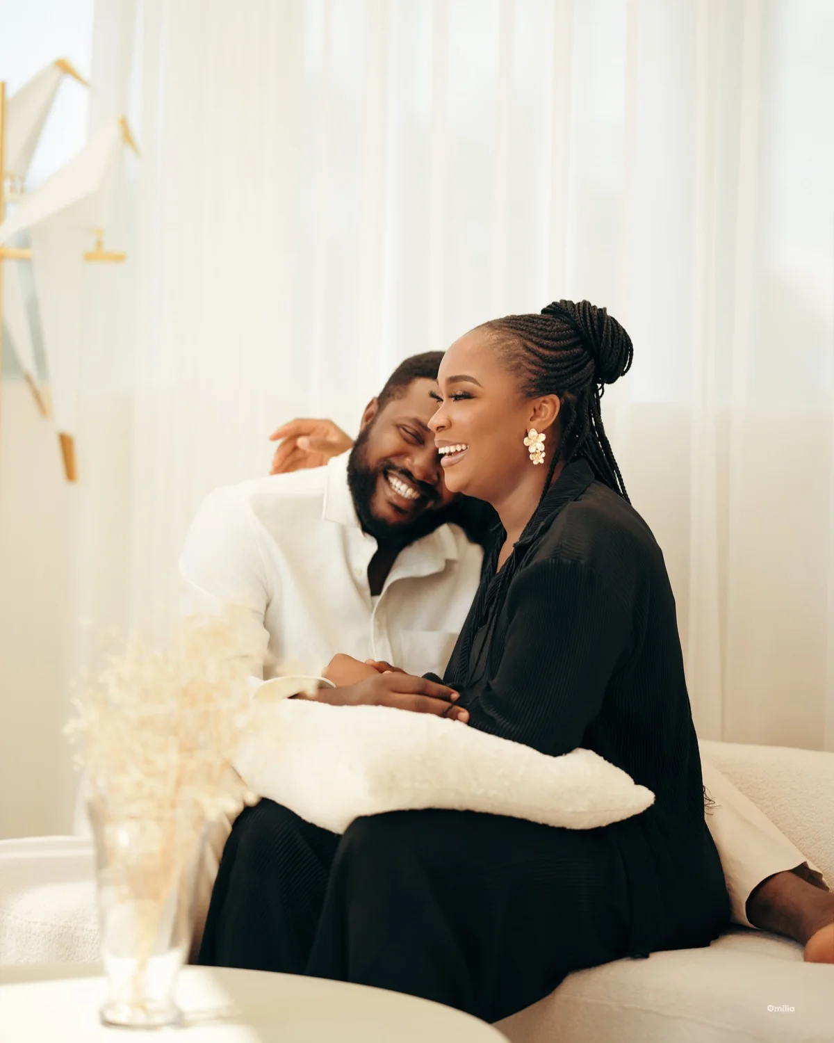 Couple laughing together on a cream sofa by sheer curtains, holding a fuzzy white pillow