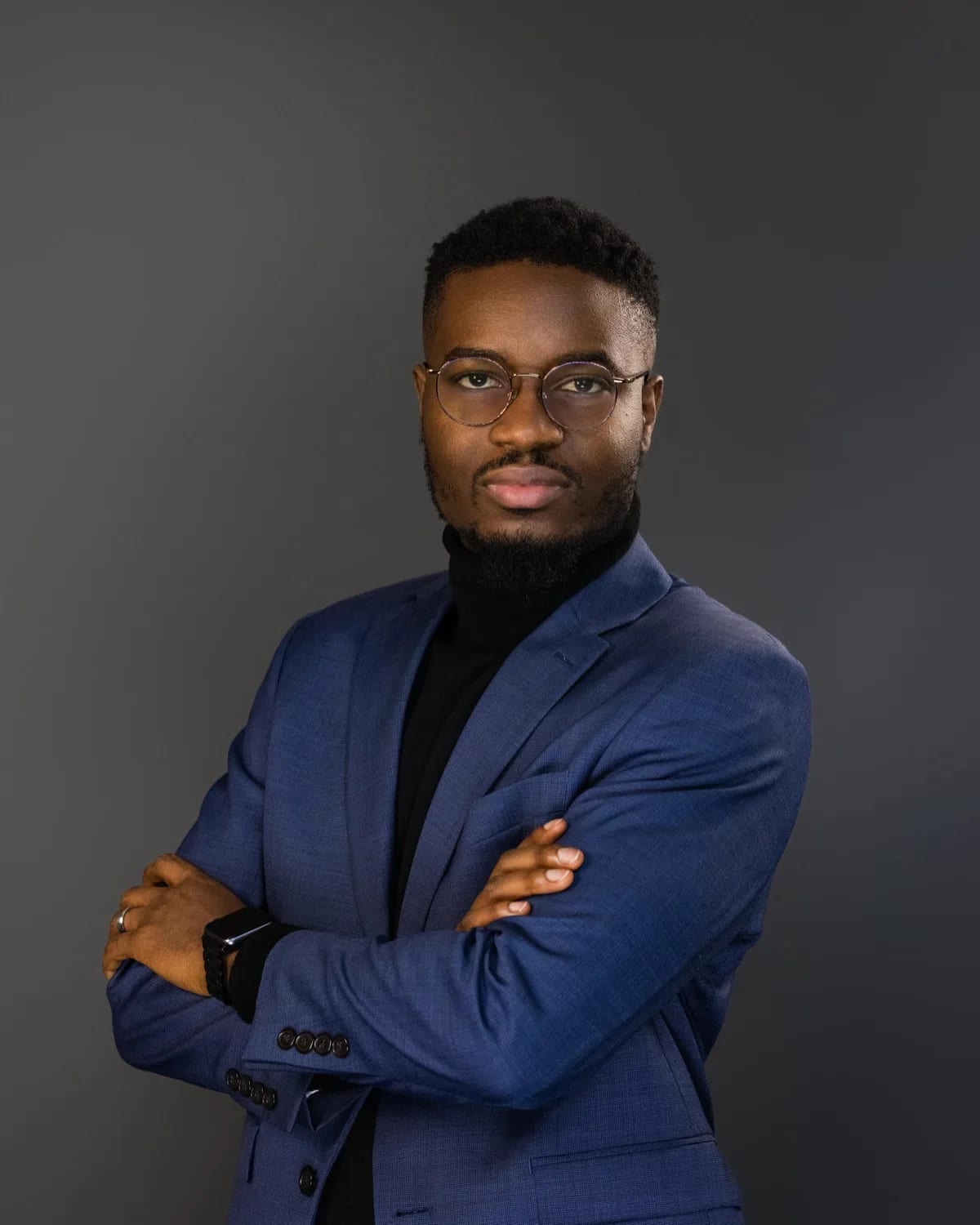 Man in blue blazer and black turtleneck with round glasses, arms crossed against gray backdrop