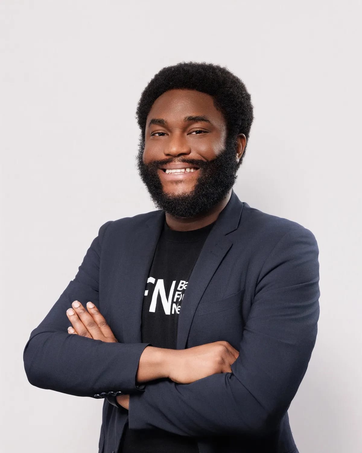 Man with full beard smiling in navy blazer over black t-shirt, arms crossed, light gray backdrop