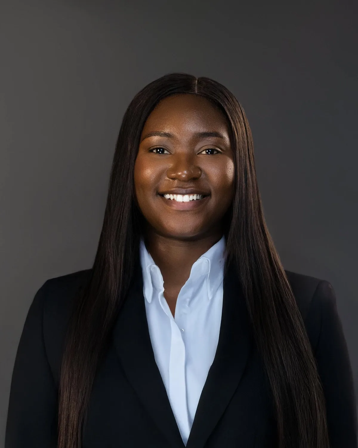Smiling woman with long straight hair in black blazer and white shirt on grey backdrop