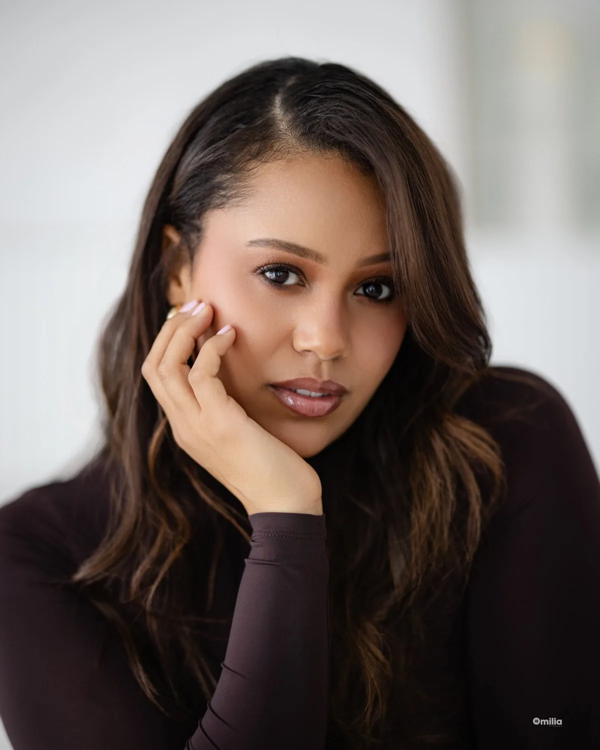Close-up of woman with long wavy hair resting hand on cheek, dark turtleneck, bright background