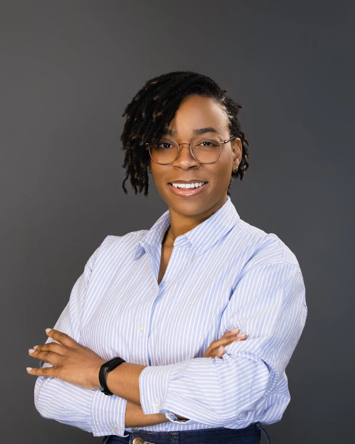 Smiling woman in glasses and blue striped shirt with arms crossed against a plain grey backdrop