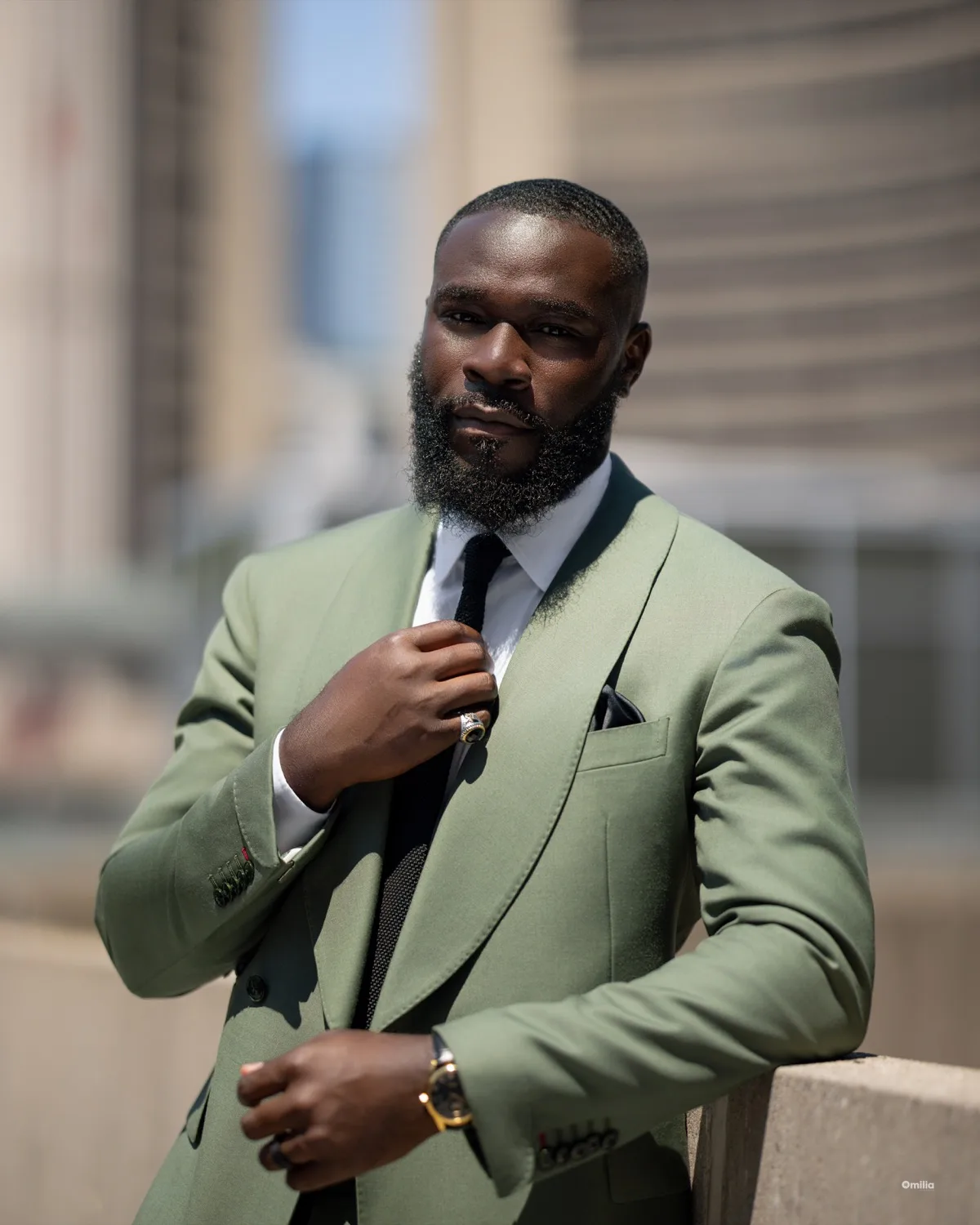 Bearded man in sage green suit adjusting his tie outdoors, leaning on a concrete ledge