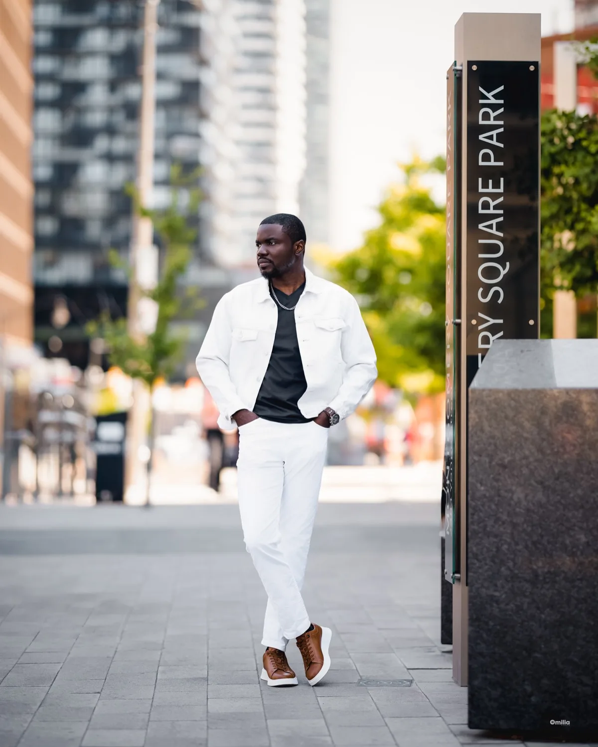 Man in white denim jacket and black tee standing on city sidewalk with blurred high-rises
