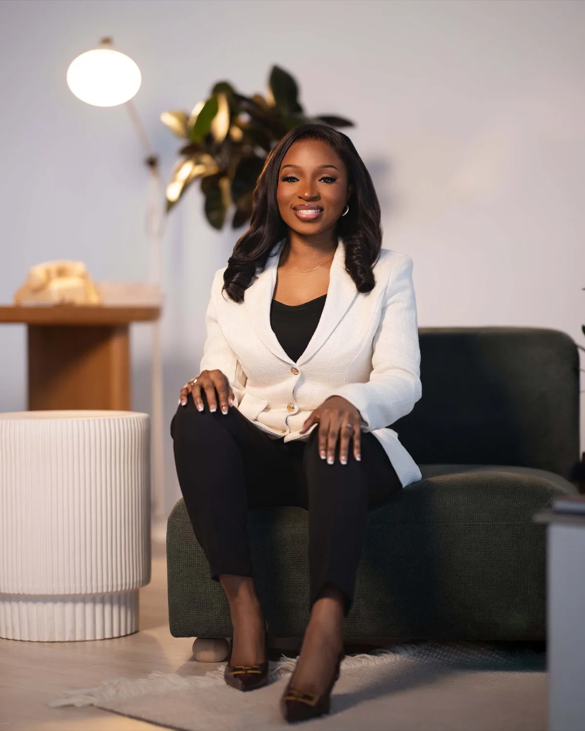 Smiling woman in cream blazer and black trousers seated on green armchair in warmly lit room