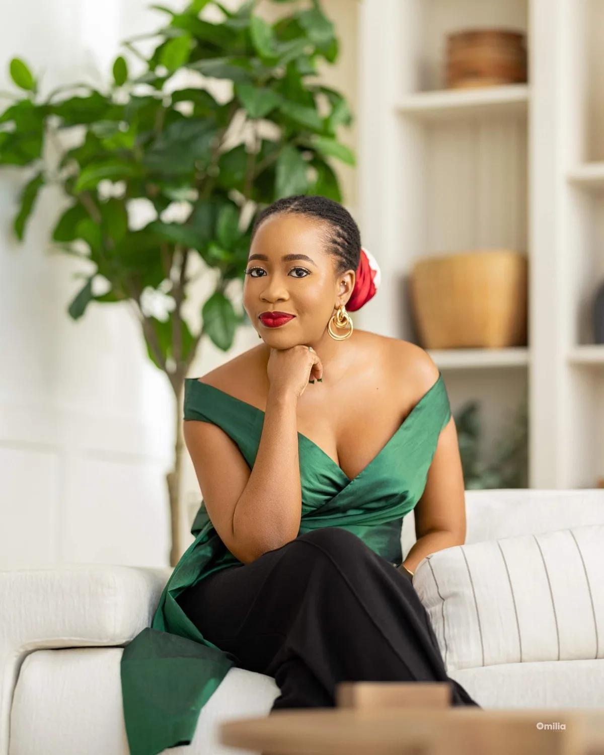 Woman in green off-shoulder top with red hair wrap, resting chin on hand on white sofa