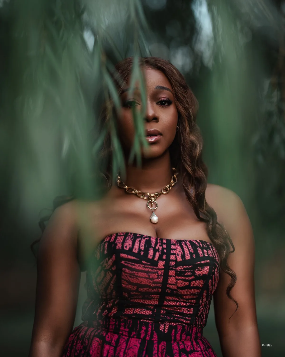 Woman in strapless pink patterned dress with gold pearl necklace framed by willow leaves outdoors
