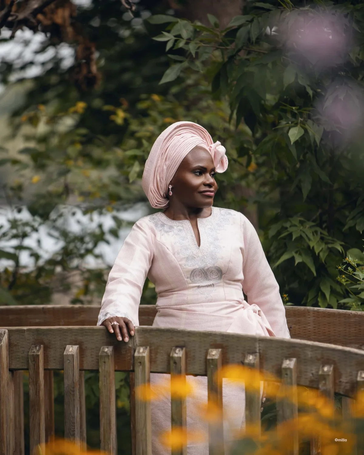 Woman in pink traditional attire and head wrap by a wooden garden railing amid greenery