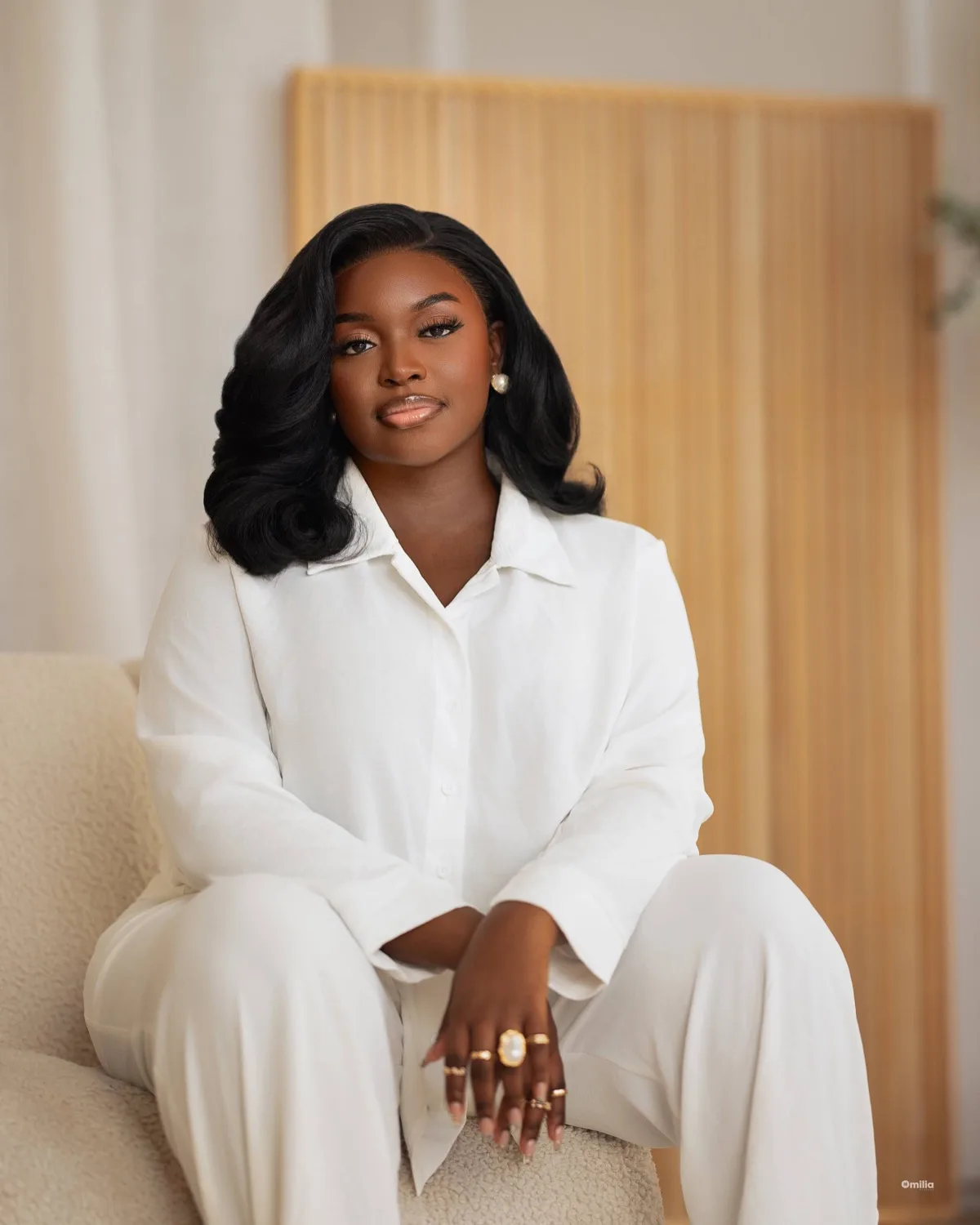 Woman in white shirt and trousers seated with clasped hands against a warm wooden slatted backdrop