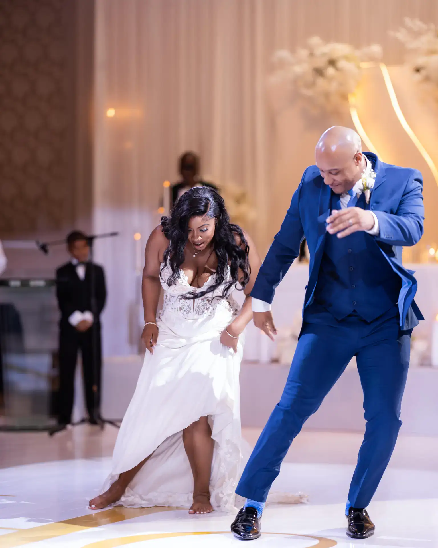 Bride lifting her gown hem while dancing barefoot beside a man in a blue three-piece suit