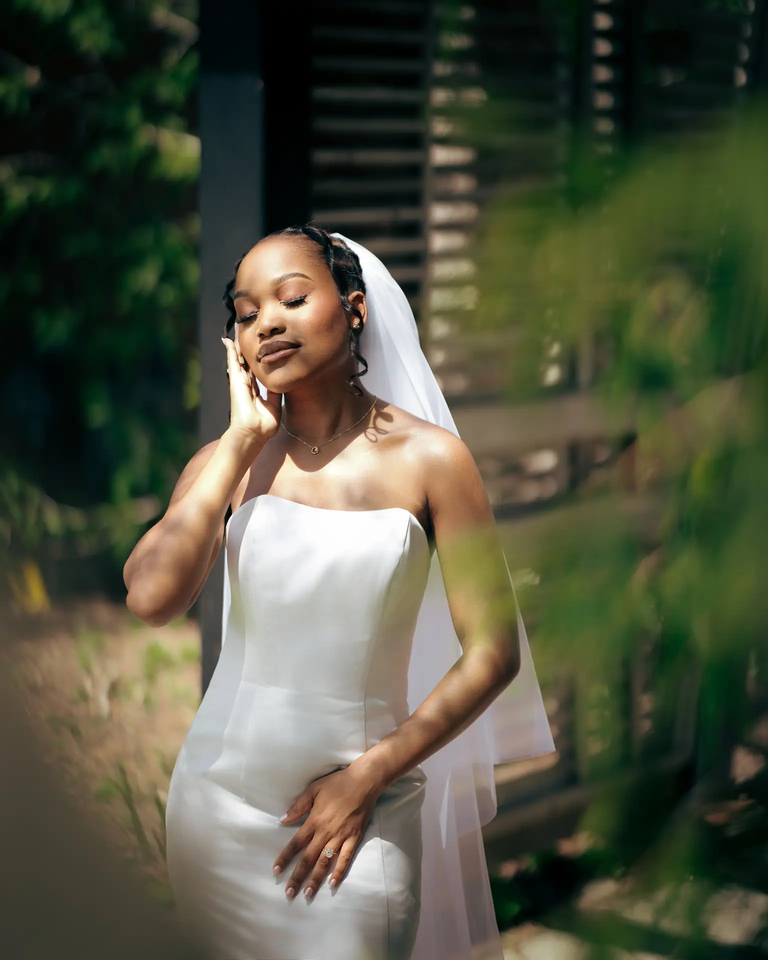 Bride in strapless gown and veil posing in dappled sunlight with hand raised to cheek