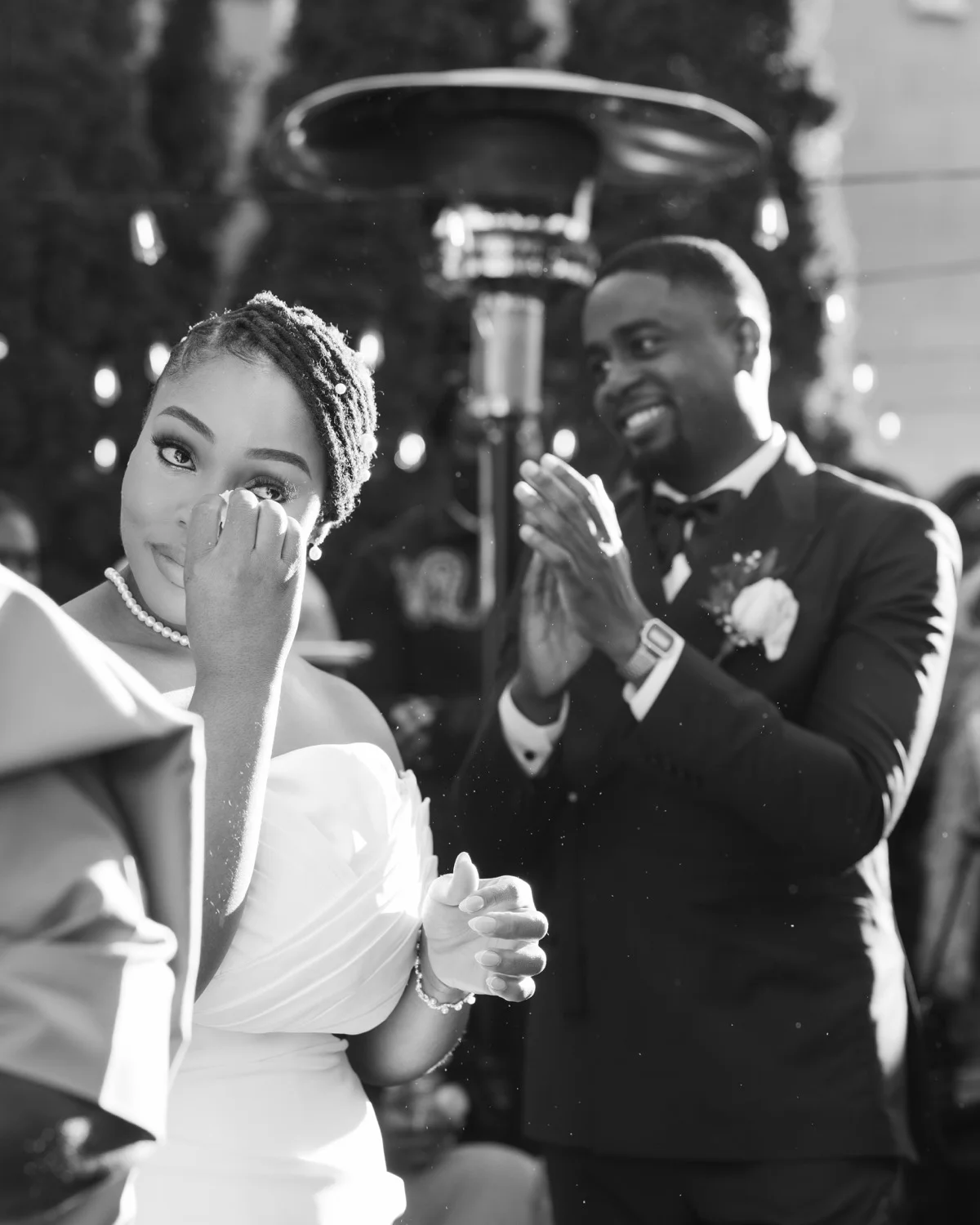 Bride wiping a tear while the groom claps behind her at an outdoor celebration