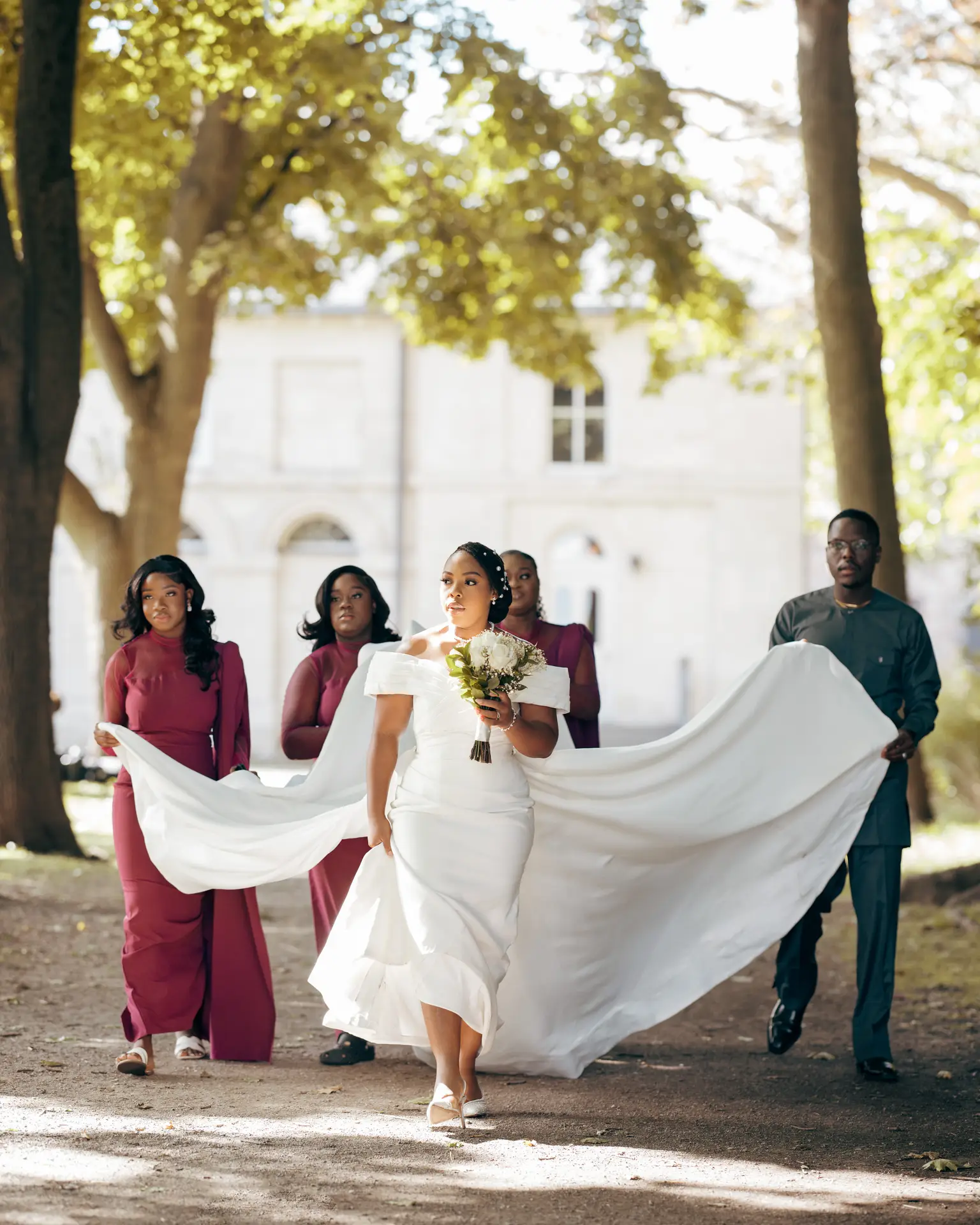 Bride holding a white bouquet as attendants fan her sweeping train along a shaded path