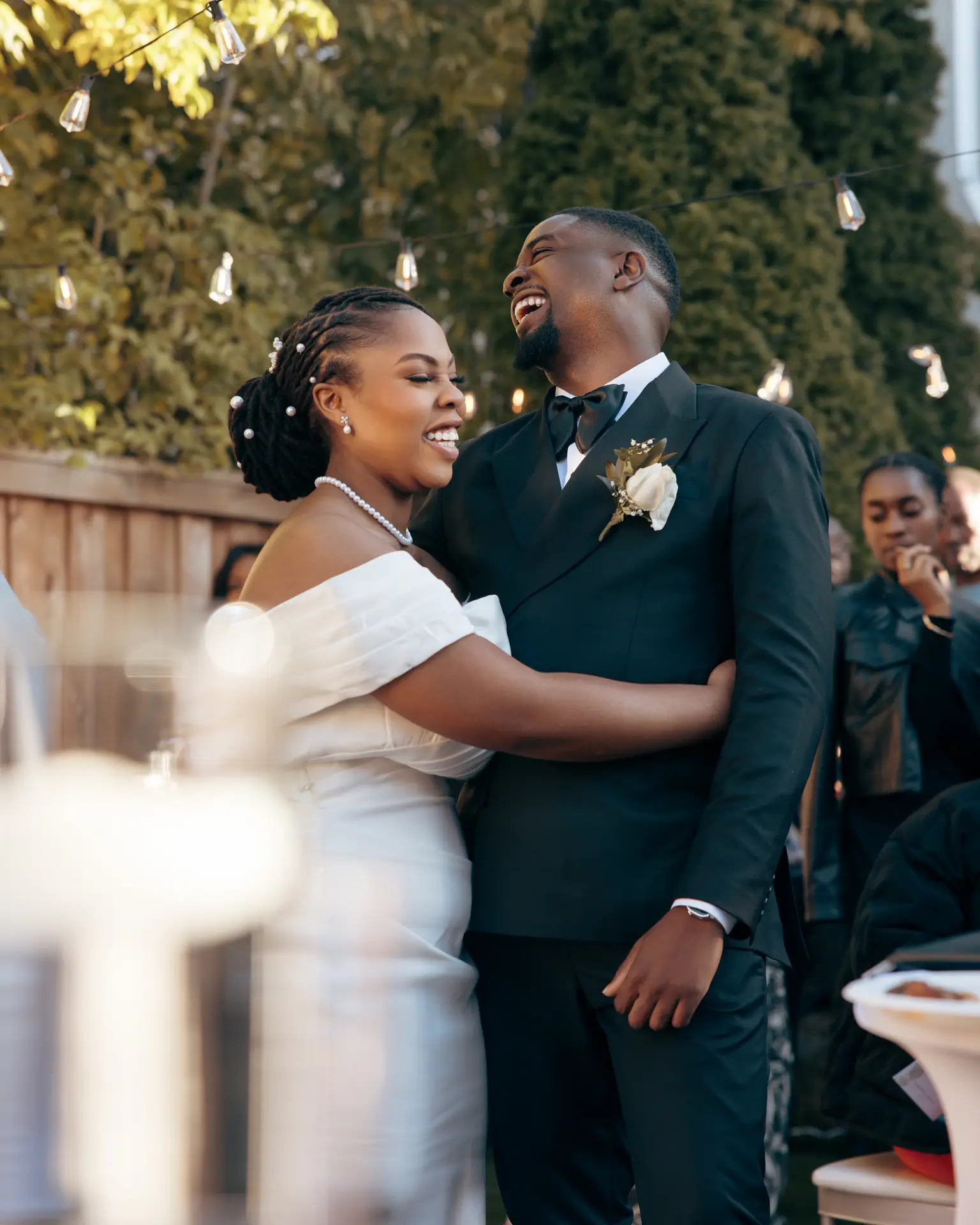 Bride and laughing groom embracing under string lights at an outdoor celebration
