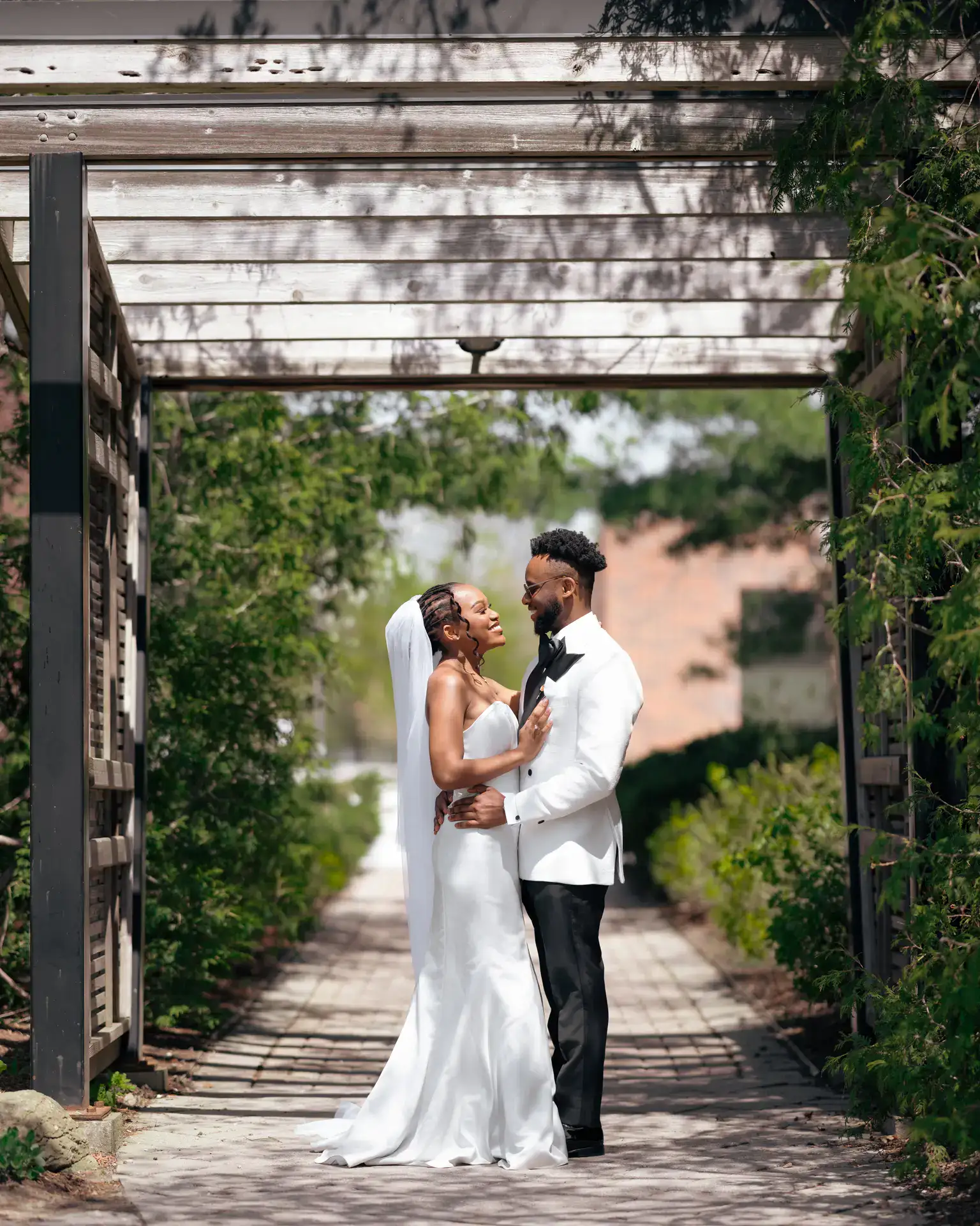 Bride and groom embracing under a wooden pergola along a garden walkway