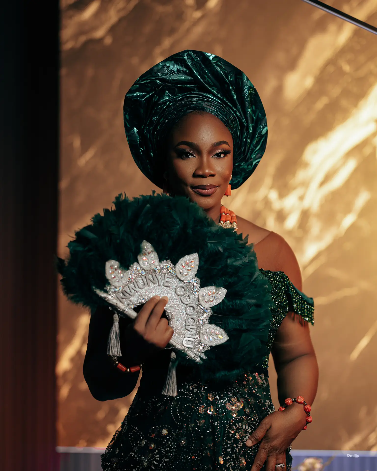 Woman in emerald gele and beaded gown holding a feathered fan against golden backdrop