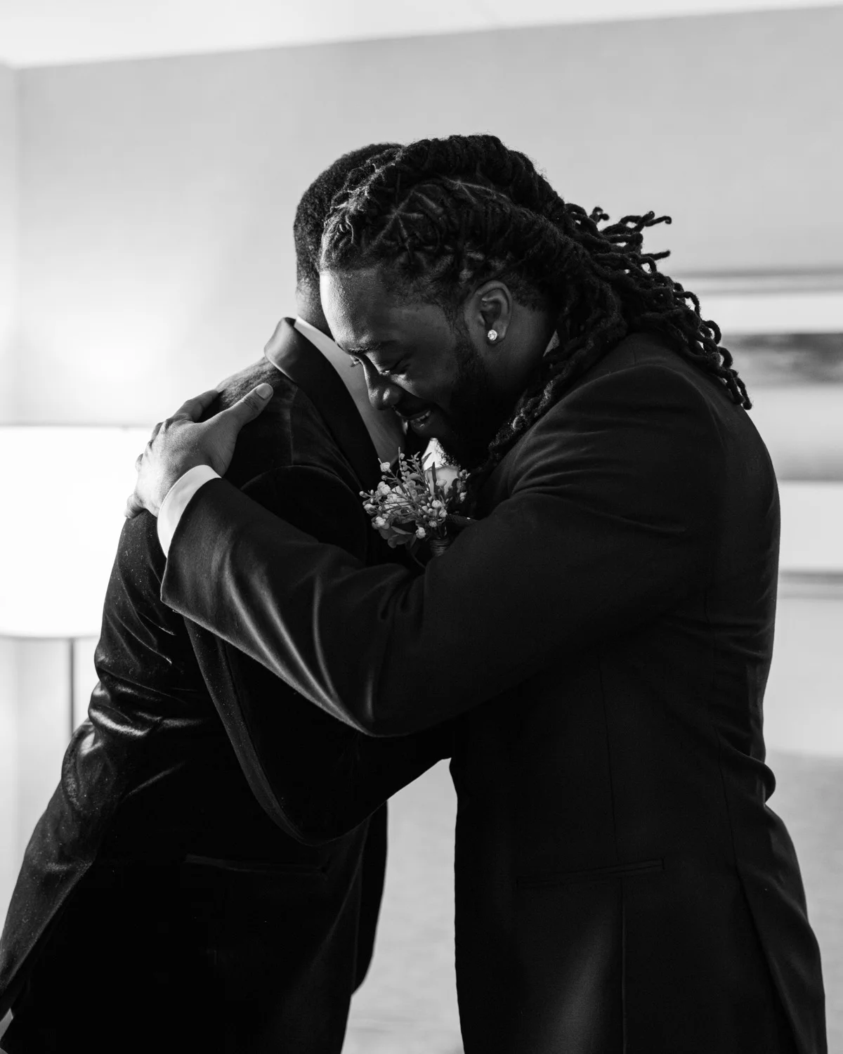 Two men in tuxedos sharing an emotional hug indoors, one wearing a boutonniere