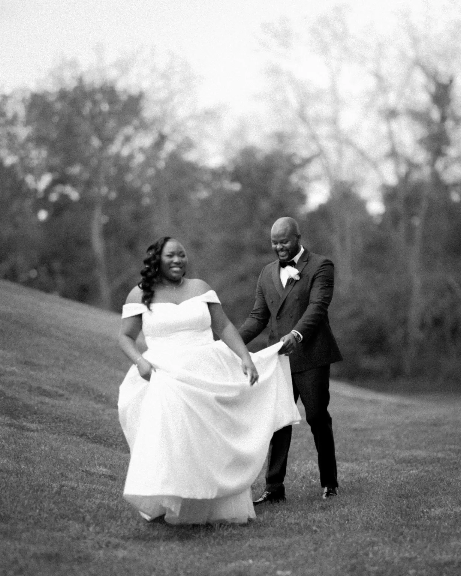 Groom carrying bride's dress train as she walks smiling across a grassy slope