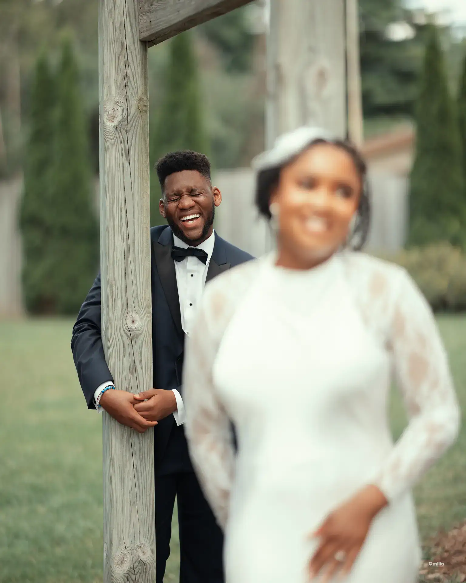 Groom laughing behind a wooden post with the bride smiling softly in the foreground