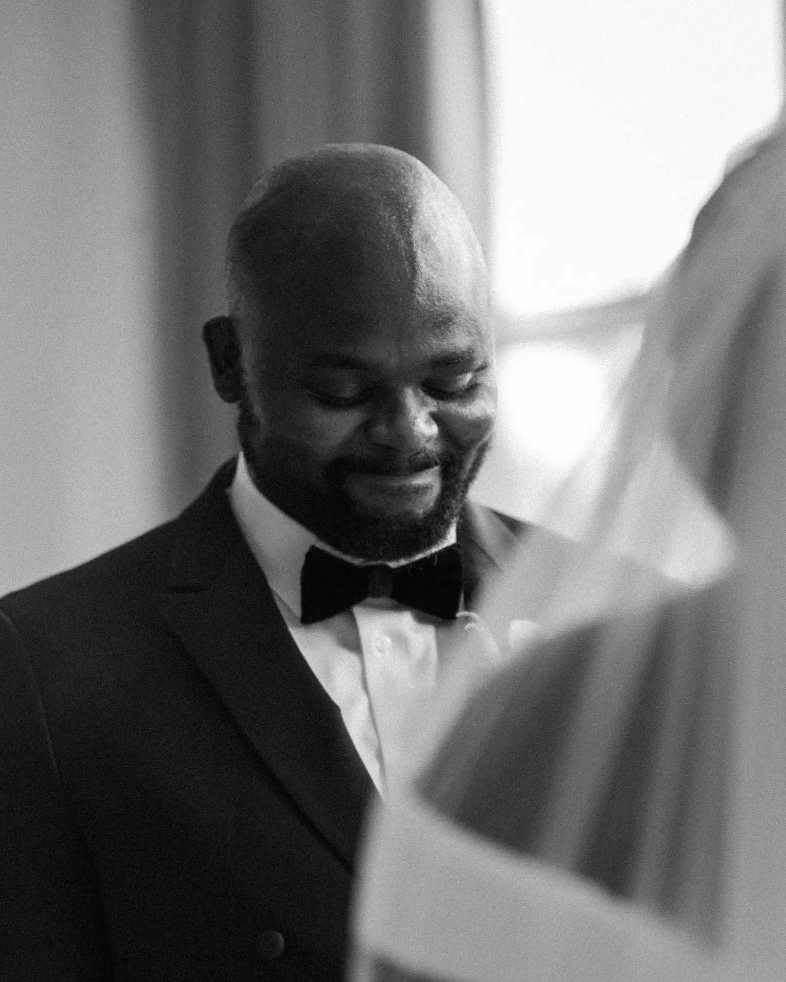 Groom in bow tie smiling softly by a window behind blurred pale fabric