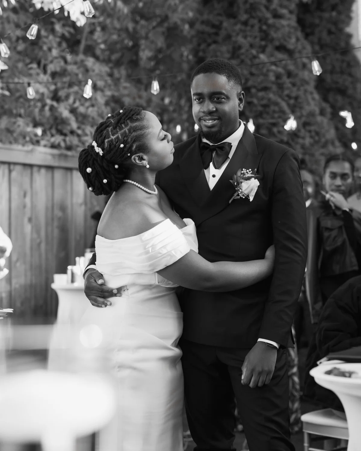 Bride with pearl-adorned hair embracing tuxedoed groom under string lights at reception