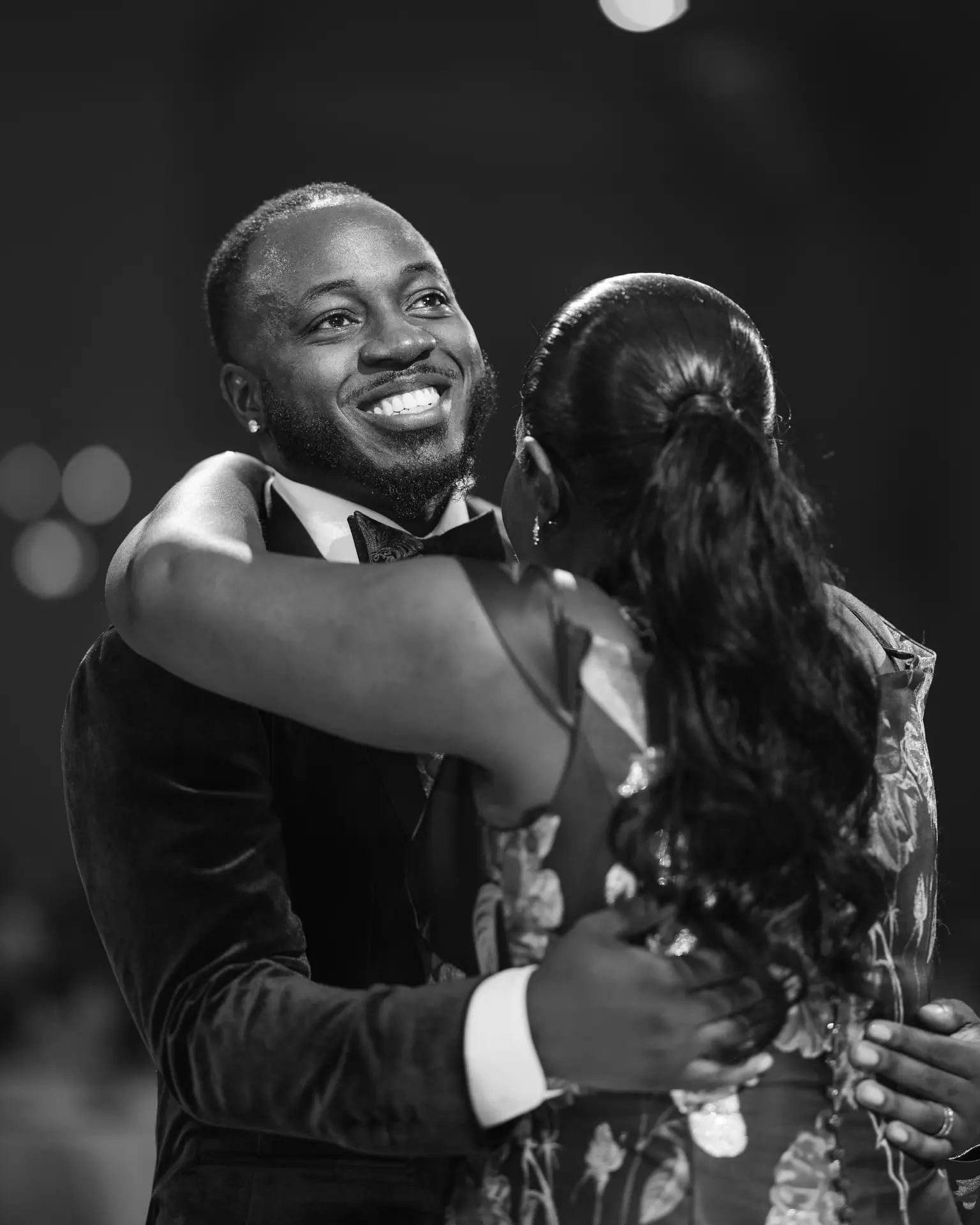 Smiling man in velvet tuxedo embracing a woman in a floral gown during a reception dance