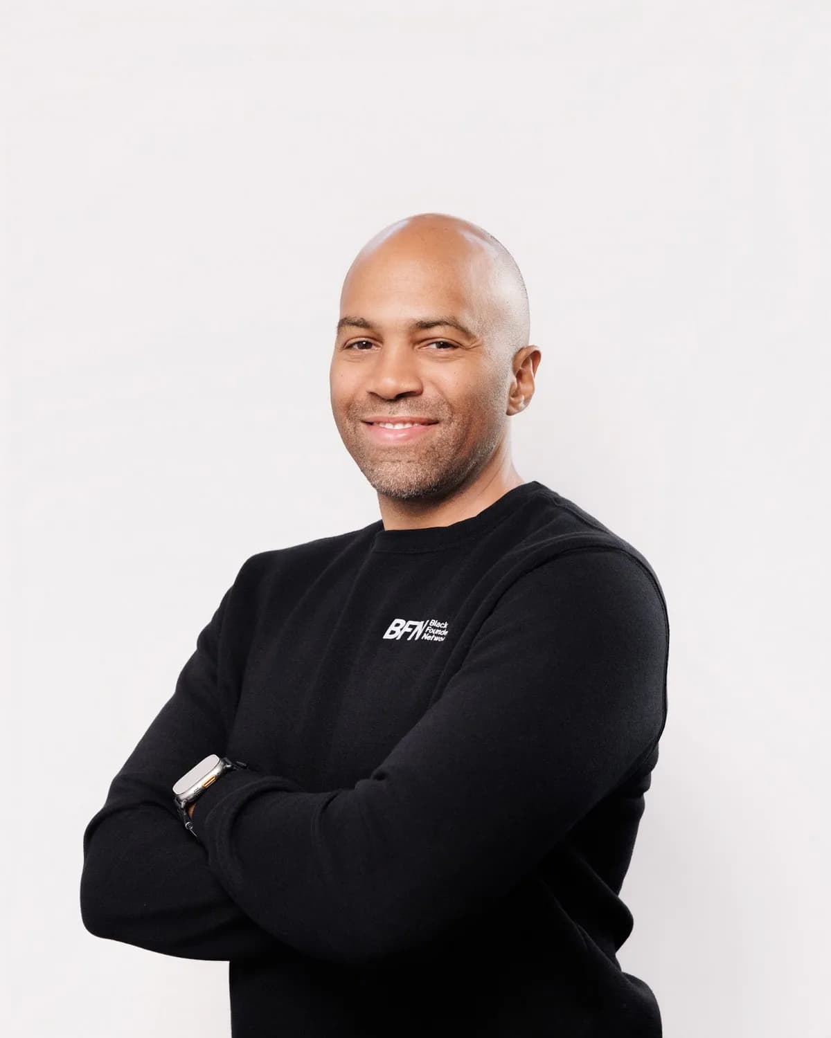 Smiling bald man with light stubble, arms crossed in black sweater against white studio backdrop