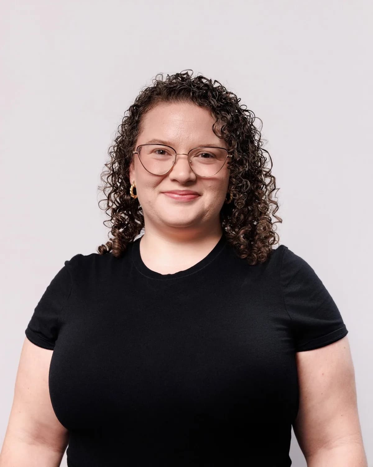 Woman with curly hair and gold cat-eye glasses in black t-shirt against light grey backdrop
