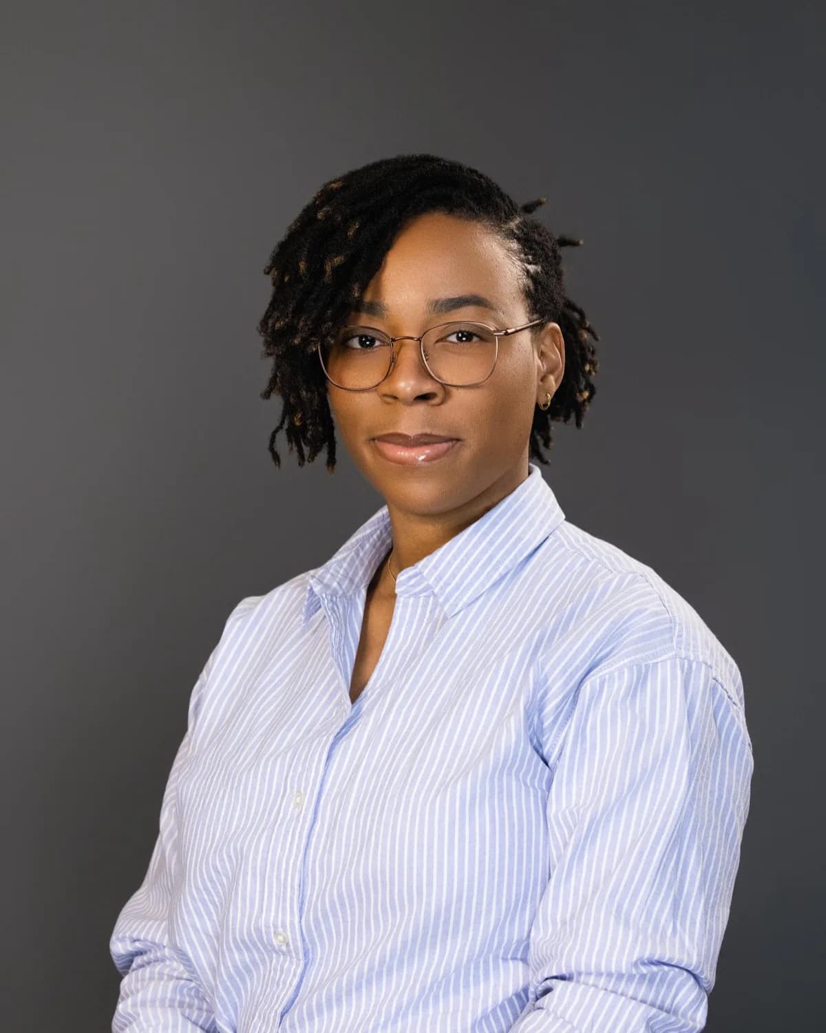 Woman with short locs and round glasses in a light blue striped shirt, grey studio backdrop